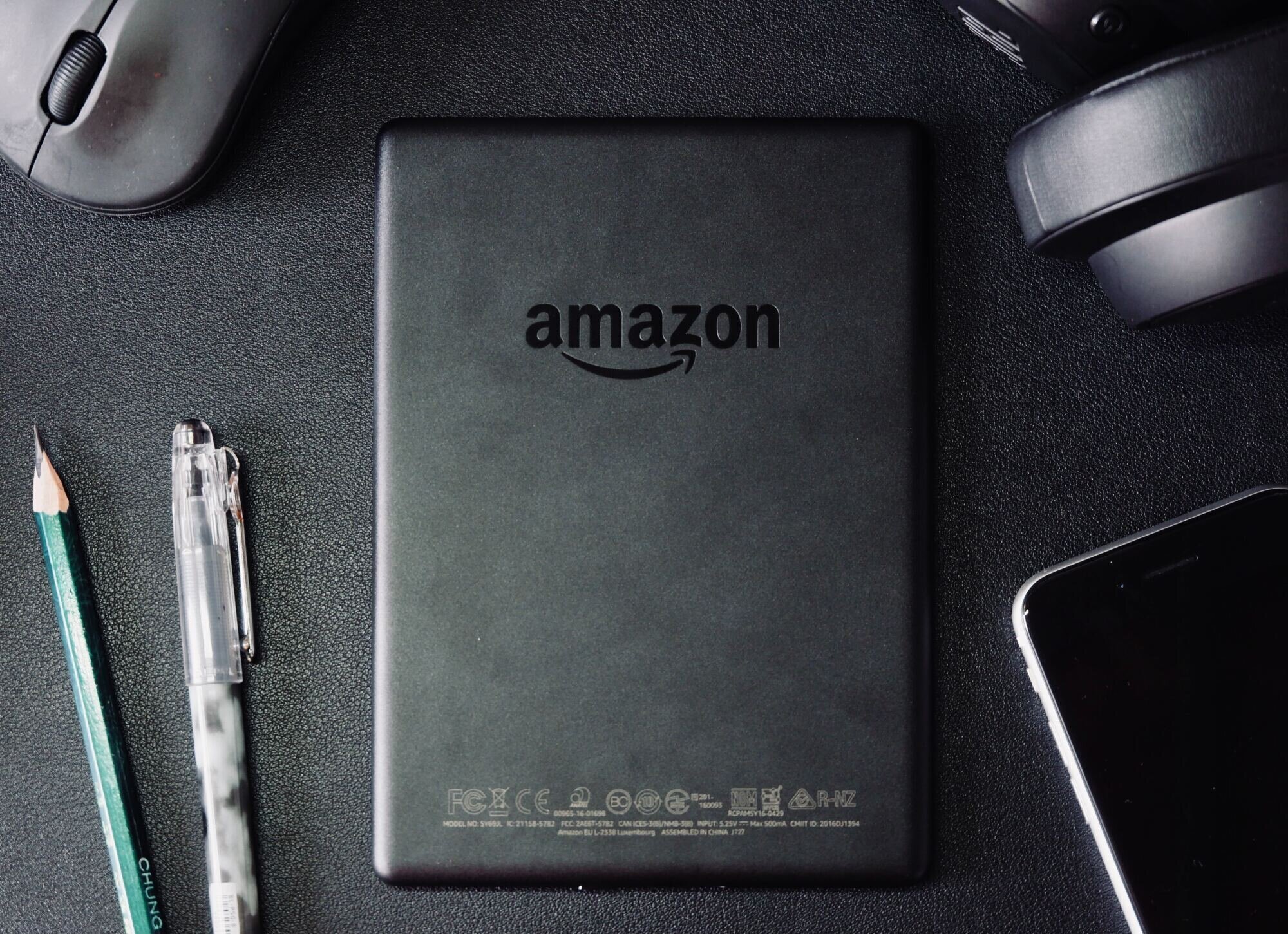 Amazon tablet back with the amazon logo, in the center of a desk mat. A mouse, headphones, pen and smartphone partially visible. All black.